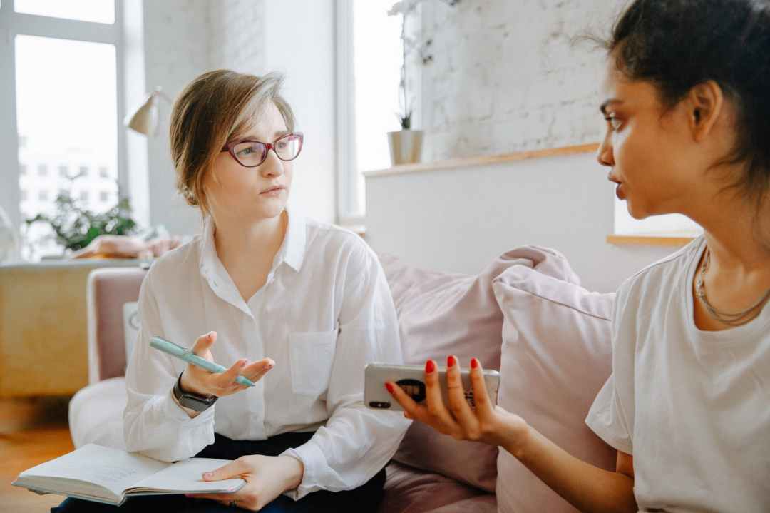 Two young women sit in their apartment, appearing to be having an upsetting conversation. They are wearing white. One wears glasses and has a pen in her hand, and the other is holding her phone. They appear frustrated.