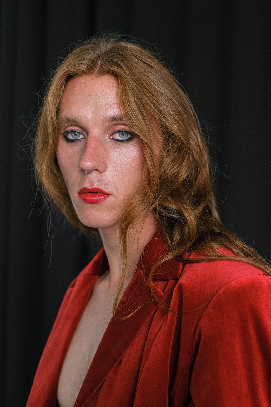 A white person with shoulder length auburn hair, under-eye liner, and red lipstick to match their top. They are are bare chested beneath the blazer and gazing dully at the camera.